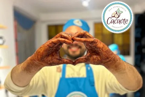 Un hombre joven con delantal y gorro azul muestra sus manos untadas de cacao y haciendo una forma de corazón.