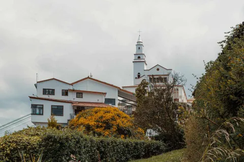 Cerro de Monserrate vista lateral de la iglesa en plano general rodeada de vegetación y árboles. El cielo gris está cubierto por nubes.