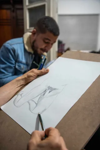  Chucuy Club con un dibujo de un pene en una hoja de papel, manos de dibujante en primer plano y de fondo un hombre jóven parece que dibujando.
