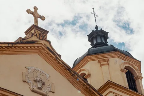 Iglesia de La Candelaria, parte superior de una de las torres de color entre amarillo y negro terminada en cúpula con cruz metálica negra mirando al cielo azul con nubes blancas.