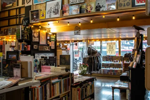 Inside La Valija de Fuego with shelves full of books, even on the walls, and a display case filled with clothing, patches, and decorative items. Outside, you can see the street in daylight.