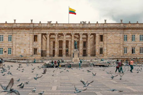 Plaza de Bolívar vista norte de a sur con vuelo de palomas, personas y una estatua de Simón Bolívar, y al fondo el Capitolio nacional donde ondea una bandera de Colombia.