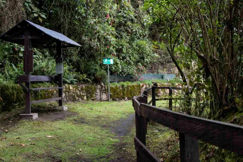  Sendero El Paramuno en curva de camino de tierra con hierba demarcado con una cerca de madera y unas rocas grandes cubiertas por musgo. Hay dos señalizaciones, una en madera, cubierta con teja para cubrirse de la lluvia y otra metálica de color verde que orienta la direccion a seguir.