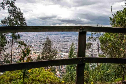 Sendero Cerro Monserrate con vista de la ciudad desde una de las cercas de madera y árboles abajo en la montaña. El cielo es gris.