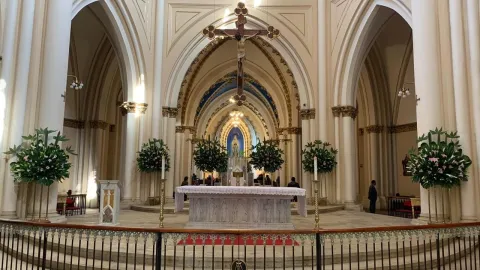 Iglesia Lourdes al interior plano general del altar con sus paredes ovaladas y un Cristo en el centro, al fondo la Virgen con personas que le oran. El altar está decorado con arreglos florales y dos velones. 