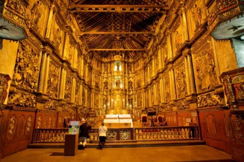  Iglesia San Francisco colonial con su interior, techos y paredes en retablos de madera dorada. Dos mujeres de rodillas oran al frente del altar blanco iluminado con varios velones. Al frente se observan figuras de varios santos iluminados con luz artificial.  