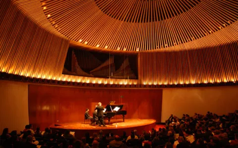 Luis Ángel Arango Library, interior concert hall with musicians on stage playing a brown grand piano. The hall, with reddish wooden walls and ceilings, is packed. 