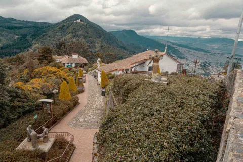 Cerro de Monserrate, sendero interno rodeado por pinos medianos y árboles en el que se ven estatuas de bronce, dos edificaciones y al fondo el cerro de Guadalupe y parte del suroccidente de la ciudad.