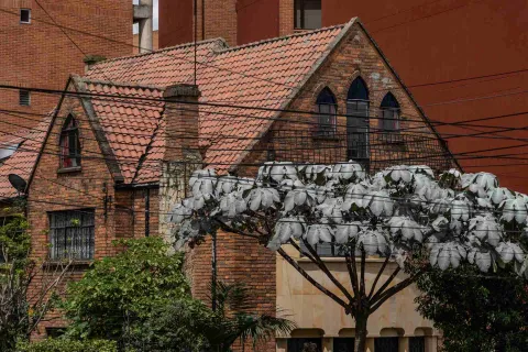 Chapinero primer plano fachada casa de tres pisos estilo inglés en ladrillo con tejas de barro, chimenea y algunos árboles al frente que la cubre. Detrás se ven altos edificios en ladrillo.