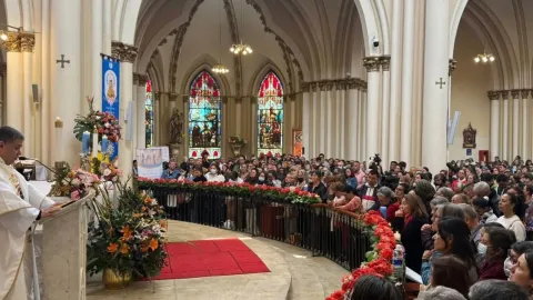 Iglesia Lourdes al interior personas cerca al altar asisten a una celebración eucarística. Un sacerdote da el sermón y habla al micrófono. 