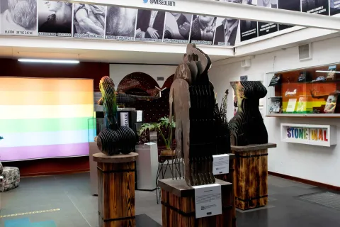 Museo Lgbti al interior salón de exhibición de obras en cobre, una con forma de falo gigante y otra un cuerpo de mujer. Hay fotografías de personalidades gay históricas y una pantalla con los colores de la bandera arcoíris lgbti.  