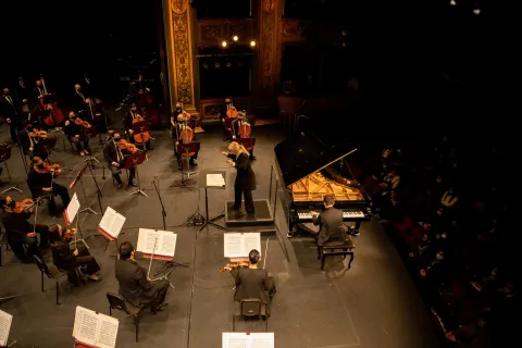 Teatro Colón, view of the stage from a balcony with musicians playing instruments and a conductor directing.