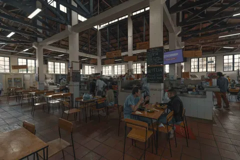 La imagen muestra el interior espacioso de una plaza de mercado o un patio de comidas. Varias personas están sentadas en mesas de madera y sillas de metal, disfrutando de sus comidas. Al fondo, se pueden ver diferentes puestos de comida con cocineros y personal atendiendo. El techo alto, con vigas de madera y metal, y las grandes ventanas, sugieren una estructura antigua y bien iluminada. Un televisor montado en la pared y letreros escritos a mano añaden detalles al ambiente.