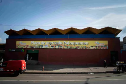 Esta imagen presenta una vista frontal más amplia de la fachada de la "Plaza de Mercado Paloquemao". El edificio de ladrillo rojo está coronado por una marquesina de techo con un diseño ondulado y escalonado en tonos amarillos y azules. Un gran letrero amarillo con el nombre del mercado y un mural ilustrado de productos frescos se extiende a lo largo de la pared. En primer plano, se ve una acera con un hidrante rojo y algunas personas caminando. Un vehículo rojo está estacionado a la izquierda.