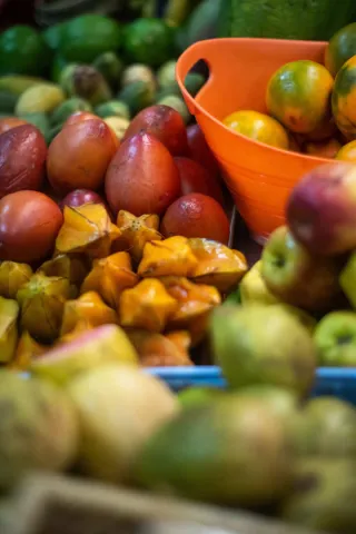 Un primer plano enfocado en una colorida variedad de frutas exóticas y frescas, dispuestas en un puesto de mercado. En el centro, se destacan varias carambolas (frutas estrella) de color amarillo brillante y frutas rojizas. A la derecha, un recipiente naranja contiene más frutas redondas. El fondo y el primer plano están suavemente desenfocados, mostrando una profusión de otras frutas verdes y amarillas, lo que sugiere una abundancia de productos.