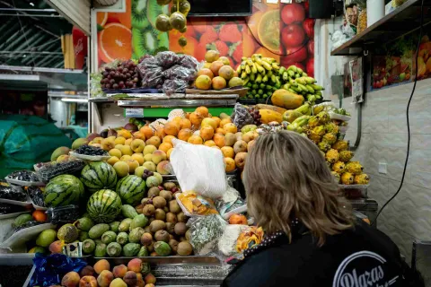 Esta imagen se centra en una vibrante exhibición de frutas frescas en un puesto de mercado. Una persona con cabello largo y rubio, vestida de negro, se encuentra de espaldas, mirando la gran variedad de frutas. Melones, uvas, plátanos, mangos, papayas y otras frutas exóticas están apiladas en abundancia. Un mural de frutas se ve en el fondo.