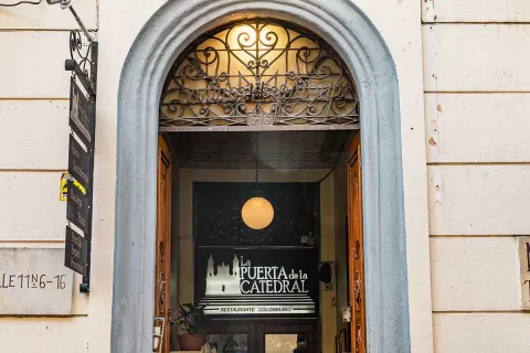 Esta imagen de cerca muestra una entrada icónica en el centro de Bogotá. Se trata de 'La Puerta de la Catedral', un restaurante ubicado en el corazón de La Candelaria. La lámpara colgante en el interior y el letrero discreto invitan a los transeúntes a descubrir un lugar lleno de historia y sabor, justo en las calles del centro histórico.