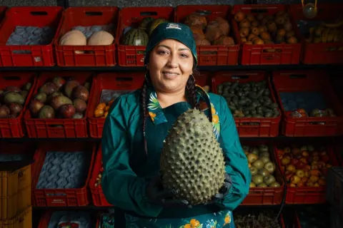 Esta fotografía de cerca muestra a una mujer sonriente en un puesto de frutas y verduras. Viste un delantal y una gorra de color verde, y sostiene en sus manos una guanábana grande y espinosa. Detrás de ella, se ven estantes llenos de cajones con diversas frutas tropicales, como melones, sandías, mangos, aguacates y cocos. La imagen resalta la frescura de los productos y la amabilidad de los vendedores, que hacen de los mercados locales un lugar vibrante y lleno de vida.