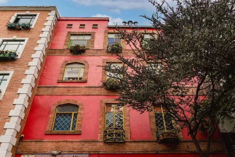 Esta foto de una fachada en La Macarena resalta la combinación de estilos y el uso del color. El edificio, con su llamativa fachada de color rojo con detalles en amarillo, se ve en contraste con un edificio de ladrillo en el lado izquierdo. Las ventanas con rejas, los balcones circulares y los adornos en la parte superior le dan un toque único y llamativo. La textura de la pintura y los detalles de la arquitectura se aprecian con claridad, lo que resalta el carácter artístico de la zona.