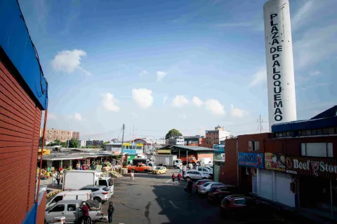 La fotografía ofrece una vista elevada de un área interior concurrida de la "Plaza de Mercado Paloquemao". Una calle asfaltada atraviesa el centro, con varios coches estacionados y personas caminando. A la izquierda, se observan puestos de mercado al aire libre con toldos. A la derecha, un edificio de ladrillo y una alta columna blanca con el texto "PLAZA DE PALOQUEMAO" escrito verticalmente. El cielo azul con algunas nubes dispersas se extiende por encima de un paisaje urbano.