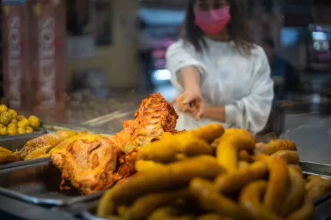 La imagen muestra una vitrina de comida de una Fritanga tradicional. En primer plano, se ven trozos de carne asada o frita, con huesos visibles, y una gran cantidad papas criollas, gallina y chunchullo. Detrás del cristal, una mujer con una mascarilla rosa y una sudadera blanca extiende la mano hacia la comida, sugiriendo que está a punto de servir o tomar un plato.