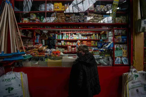  Esta imagen muestra el interior de una pequeña tienda. Una persona con abrigo oscuro está de espaldas, mirando los productos. El puesto está repleto de estantes con una variedad de artículos, incluyendo latas, paquetes de alimentos, escobas y bolsas grandes de azúcar o granos en la parte inferior. La iluminación superior se refleja en una ventana, revelando más productos apilados.