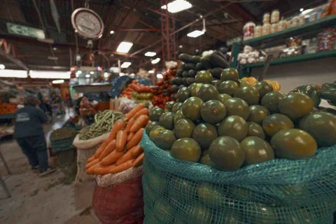 En esta imagen, un primer plano enfocado en grandes sacos de frutas cítricas verdes domina el lado derecho. Detrás y a la izquierda, se ven sacos de zanahorias, calabacines y otros vegetales apilados. Al fondo, se distingue el ambiente bullicioso de un mercado, con una persona caminando y estantes llenos de productos. Una báscula circular se ve desenfocada en la parte superior izquierda.