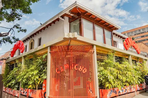  La imagen presenta la fachada de Camacho, un restaurante con un diseño llamativo y contemporáneo. El edificio, de color blanco y con toques de madera, se destaca por su estructura en el primer piso, construida con tablillas de color naranja vibrante. Este elemento geométrico y audaz crea un contraste interesante con la arquitectura circundante. La vegetación abundante a lo largo de la fachada aporta frescura y vida al espacio.
