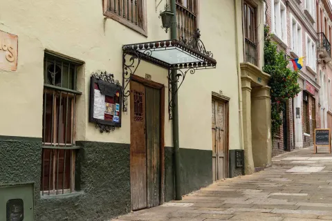 La fotografía muestra una calle adoquinada en La Candelaria. El enfoque principal está en un edificio de fachada ocre con un zócalo de color verde oscuro, que alberga lo que es una cafeteria. Una puerta de madera rústica y una ventana con rejas de hierro forjado resaltan el estilo colonial del lugar. 