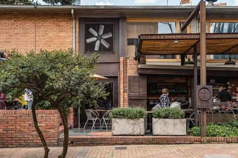 The building, with its brick walls and wood details, features a large window with a ceiling fan that adds a distinctive touch. An outdoor terrace, bordered by concrete planters filled with greenery, invites diners to enjoy the fresh air. The sidewalk bricks and outdoor tables contribute to the relaxed and modern atmosphere.