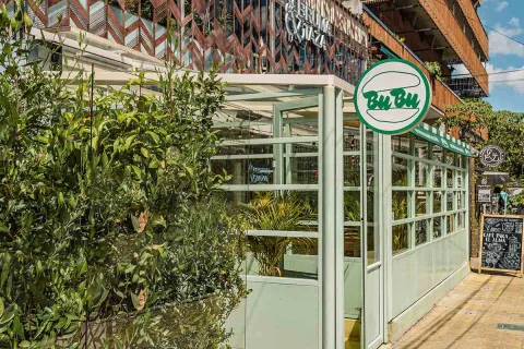Esta foto de cerca muestra una parte de un café en el Parque de la 93. La estructura, que parece ser una extensión de un edificio, está hecha de vidrio y marcos de color verde claro, lo que le da un aspecto moderno y limpio. Un letrero redondo con el nombre "Bu Bu" y un icono de una hamburguesa de estilo minimalista cuelga en la parte superior. Alrededor de la estructura de vidrio, plantas verdes aportan frescura y privacidad.