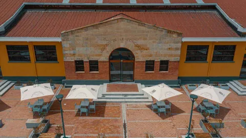 Esta vista aérea del "Mercado de la Concordia" ofrece una perspectiva única de su arquitectura y entorno. El edificio, con su tejado de terracota y sus paredes amarillas, se destaca en el centro de la imagen. La plaza exterior, con su pavimento de ladrillo y sus mesas y sillas bajo sombrillas blancas, se extiende por delante de la entrada principal. La vista desde arriba permite apreciar la armonía del diseño, que se integra perfectamente con el paisaje urbano.