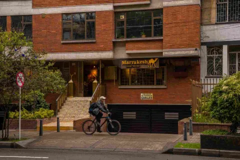 La fotografía muestra la entrada de 'Marrakech', un restaurante con una fachada de ladrillo rojo en el Park Way. La puerta principal está flanqueada por una escalera de azulejos y barandillas doradas, lo que le da un toque exótico. El letrero del restaurante, con el nombre en letras árabes y la silueta de un camello, evoca la cultura de Marruecos. 
