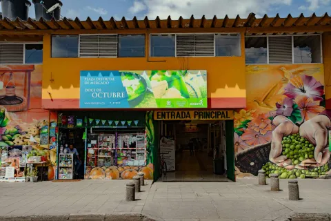  Esta fotografía presenta la fachada exterior de la "Plaza Distrital de Mercado Doce de Octubre". El edificio es de color amarillo brillante y cuenta con grandes murales vibrantes en ambos lados de la entrada principal, que representan frutas, verduras y manos sosteniendo guisantes. Un gran letrero azul con información del mercado se encuentra sobre la entrada. La "ENTRADA PRINCIPAL" está claramente marcada y abierta, revelando un pasillo interior.