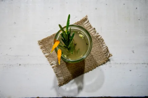 Esta es una vista cenital de un vaso pequeño con una bebida de color verde pálido, decorada con una ramita de romero y dos pétalos de naranja o cáscaras en forma de luna. El vaso está colocado sobre un pequeño trozo de arpillera cuadrada, que a su vez se encuentra sobre una superficie de madera blanca. La iluminación crea una sombra suave debajo del vaso y el posavasos.