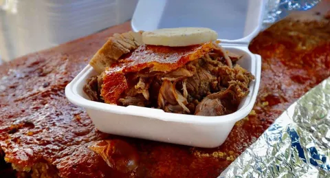 Esta fotografía de cerca se enfoca en un plato de lechona servido en un recipiente blanco de icopor. La lechona, con su carne jugosa y tierna, está coronada con un trozo de la crujiente piel de cerdo y un pedazo de arepa. La textura de la carne desmenuzada y el color dorado de la piel son los protagonistas de la imagen, que evoca el sabor y la tradición de este plato típico colombiano. En el fondo, se ve el resto de la lechona entera, lista para ser disfrutada.