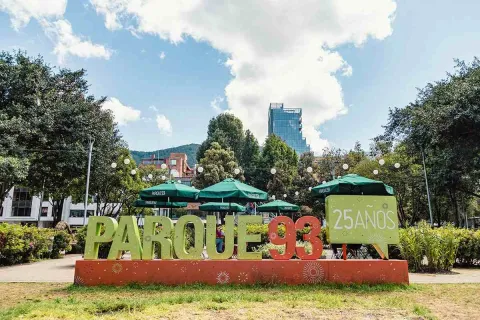 La imagen muestra una toma frontal del letrero icónico del Parque de la 93. La estructura, que celebra los "25 AÑOS" del parque, es una escultura de color naranja con el texto "PARQUE 93" en letras grandes de color verde. Detrás del letrero, se pueden ver las sombrillas de los cafés y restaurantes que rodean el parque, así como los altos árboles y un rascacielos en el fondo. El cielo azul y el clima soleado hacen que el ambiente sea vibrante y lleno de vida.