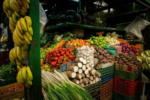 Un primer plano detallado de un puesto de frutas y verduras en el mercado, mostrando una impresionante variedad y abundancia de productos. Grandes racimos de plátanos amarillos cuelgan en el lado izquierdo. El puesto está densamente lleno de pimientos rojos, tomates pequeños, cebollas rojas, zanahorias, raíces de yuca apiladas, patatas, lechugas y otras hortalizas. Los productos están dispuestos en cestas y cajas, creando una explosión de colores y texturas.