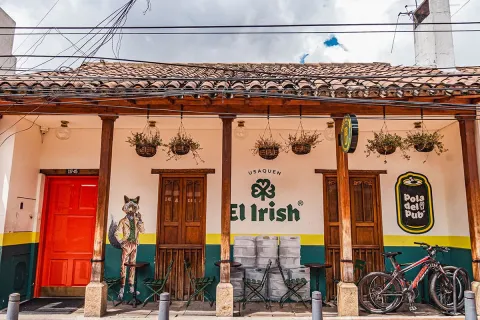 Entrada del pub “El Irish” en Usaquén, decorada con un mural artístico de un zorro elegante y un diseño que mezcla tradición e identidad irlandesa. Su fachada blanca con tejas de barro, mesas en la parte exterior y bicicletas estacionadas transmiten un ambiente bohemio y relajado.