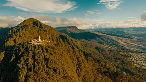 Guadalupe Hill, seen from afar atop the lush green mountain peak, offers a distant view of Bogotá. The sky is blue, dotted with white clouds.