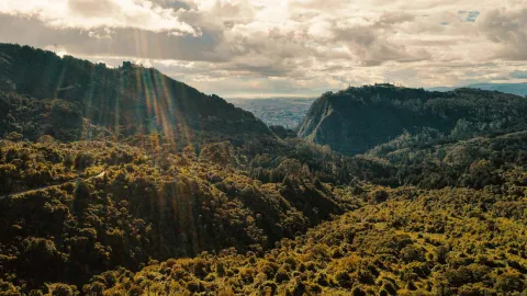 Guadalupe Hill surrounded by lush green mountains, with Bogotá visible in the distance. The sky is blue with white clouds and a rainbow.