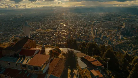 Guadalupe Church from above and the back of the white statue, surrounded by green trees, with Bogotá visible in the distance with a blue sky and white clouds.