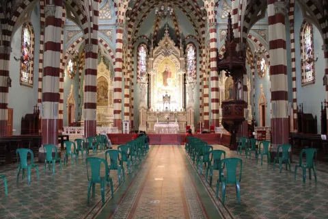 Santuario Nuestra Señora del Carmen en el interior con luz artificial destacan las columnas con franjas horizontales de color rojo y blanco, el púlpito en madera y al fondo el altar con una luz brillante sobre una imagen de la Virgen. Al centro de la iglesia hay cuatro filas de plástico vacías y dispuestas para los feligreses.
