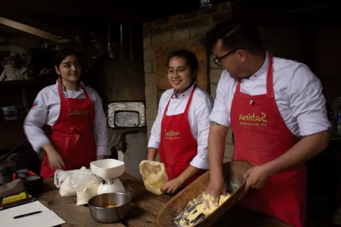 Dos mujeres jóvenes y un hombre jóven amasan amasijos en una cocina artesanal.