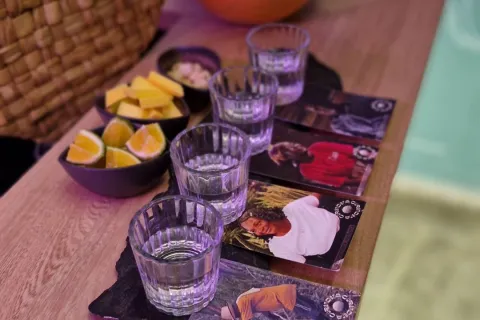 Cuatro copas pequeñas con viche sobre sendos portavasos con fotografías de personas de raza negra. En la mesa hay unas vasijas con limón, piña y maní.
