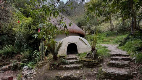 Primer plano de Maloca rodeada de árboles y vegetación. Hay una especie de gruta al frente y camino piedra alrededor.