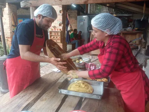  Un hombre y una mujer jóvenes cocinan con delantal y cofias unos amasijos. La cocina es artesanal.