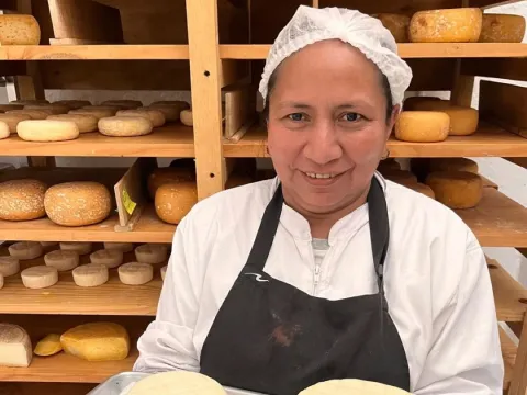 Mujer joven de tez canela sonriente con cofia delante de una estantería de madera con quesos.