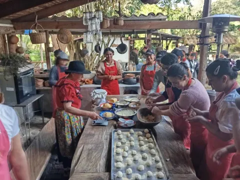  Mujer adulta con sombrero y vestido campesino enseña a cocinar a varias personas jóvenes en una concina artesanal. Hay una mesa con varios amasijos y harinas.