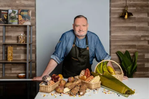 Chef Carlos Gavíria sonríe a la cámara, lleva camisa azul y delantal negro. Delante se observa una mesa con ingredientes como plátanos, papas, mazorcas y yucas. 